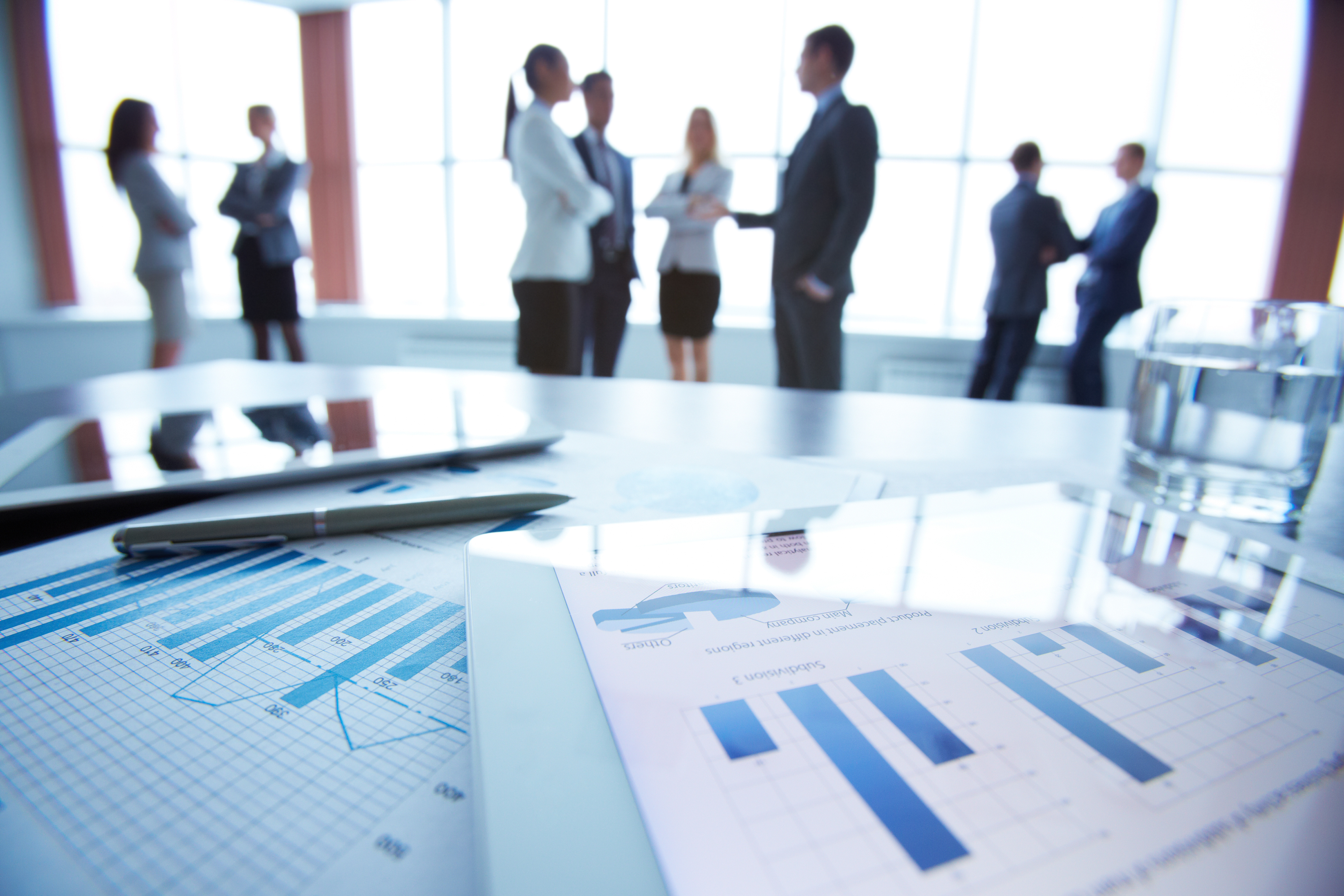 Close-up of business electronic and paper documents on the desk, office workers interacting in the background